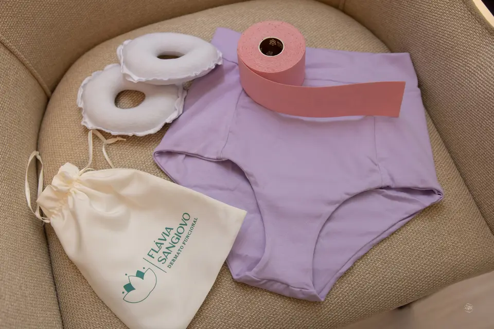 Sobre uma poltrona: rolo de taping rosa, calcinha lilás de cintura alta, duas almofadas em formato de anel e uma bolsinha de tecido com a marca Flávia Sangiovo